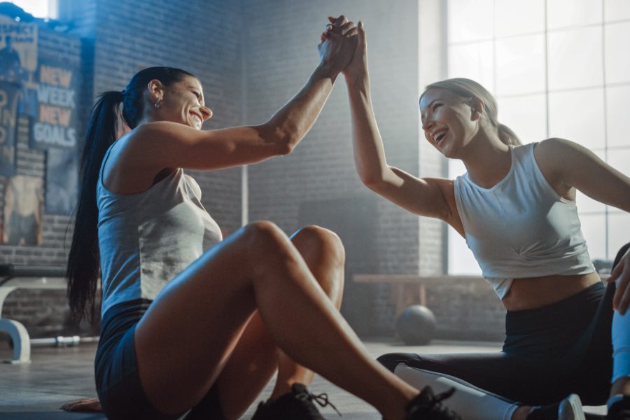 Two Beautiful Fit Athletic Girls Sit on a Floor of Industrial Loft Gym. They're Happy with their Training Program and Successfully Give a High Five.