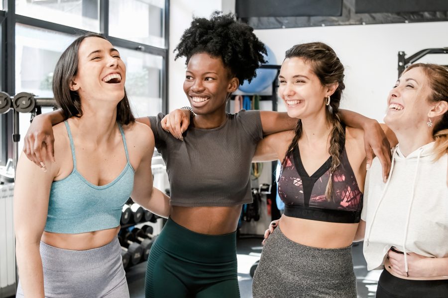 Group of happy young multiracial female friends in activewear smiling and laughing while hugging each other during break from workout in gym together